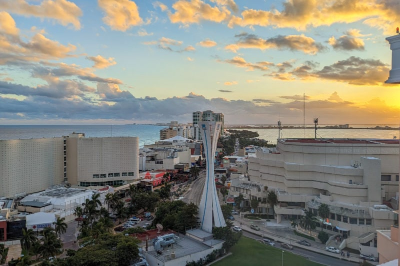 view of the hotel strip in cancun through a glass window at sunsey with the clouds in the sky turning golden. there is a long straight road with skyscrapers and large resorts on either side.