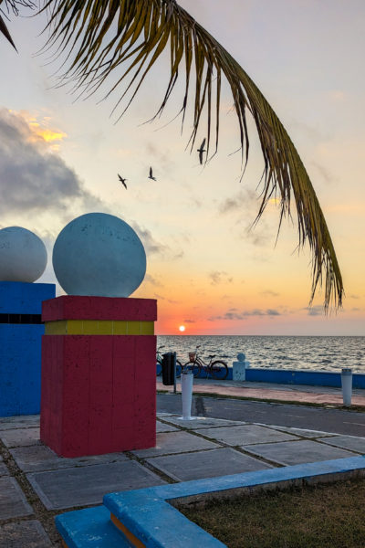 section of a paved esplanade at sunset with one red pillar and one blue pillar both topped with white stone balls. the sun is setting over the sea with a silhouetted palm leaf in the foreground. The malecon in Campeche Mexico.