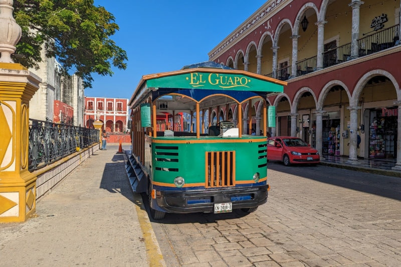 small green old fashioned tram with wooden trim and th ewords El Guapo printed at the top parked in a grey flagstone street with a park on one side and a red building with white arches on the right in Campeche main square