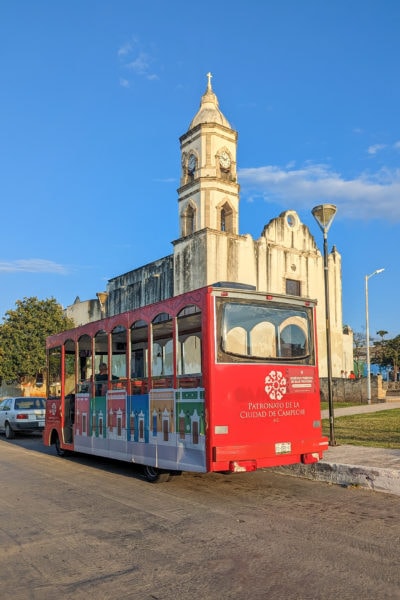 small red tram in the road in front of a white church with an arched roof and one tower against a blue sky - things to do in campeche mexico