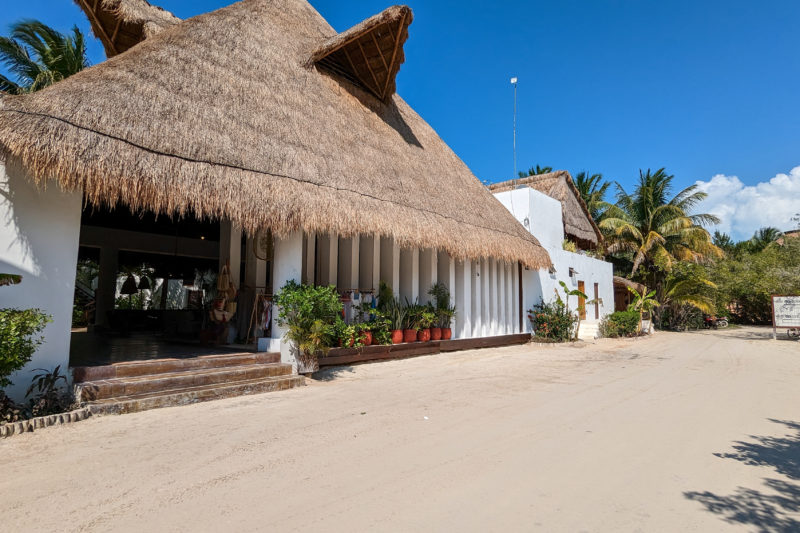 exterior of a hotel with whitewashed walls and a pointed grass roof next to a sandy road wtih clear blue sky overhead. best hotels in holbox.