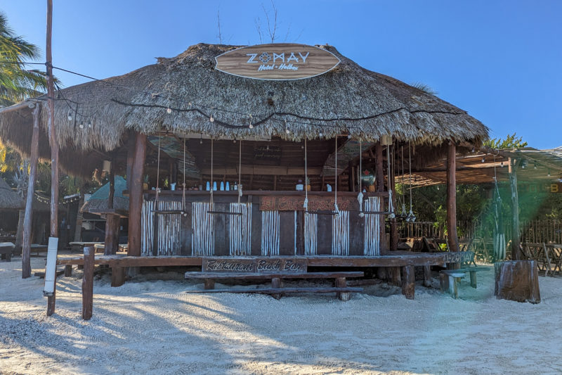 open sided wooden bar building with a row of swing seats at the bar and a grass roof with a white sandy beach in front. the sign says Zomay Hotel Holbox.