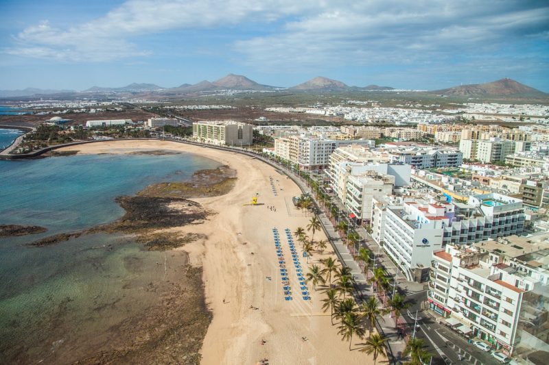 aerial shot of Arrecife city with a large and wide golden sand beach and the blue sea lined with high rise white buidlings with several mountains in the distancea and blue sky above