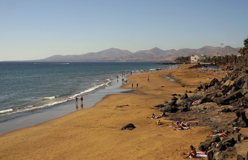 aerial shot of a golden sandy beach with a few rocks on a warm hazy day with blue sea on the left and the edge of a town on the right and low brown mountains in the distance - best areas to stay in lanzarote