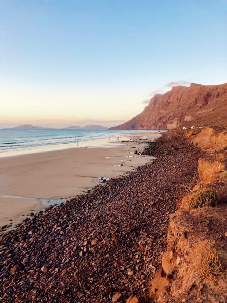 portrait shot of a beach with golden sand and orange shingle overlooked by tall rust coloured cliffs with blue sea and some low mountains visible in the far distance, taken just before sunset with a pinkish light. Famara Beach is one of the best areas to stay in Lanzarote. 