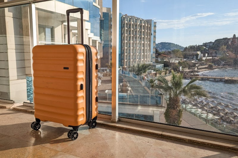 mustard yellow hard shell suitcase with a black handle on a hotel balcony next to a glass panel with a view of the ocean and marina out of focus behind. Antler Clifton Review. 