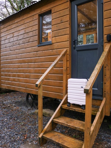 pale pink hard shell cabin sized suitcase on the steps of a small wooden cabin with a grey door