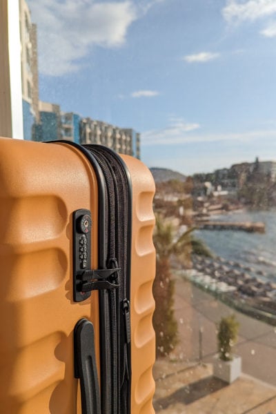 close up of the side of a mustard yellow hard shell suitcase with a black handle on a hotel balcony next to a glass panel with a view of the ocean and marina out of focus behind