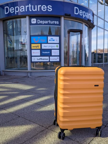 mustard yellow hard shell suitcase with a black handle on concrete floor outside a glass fronted airport building with a blue departures sign above the door