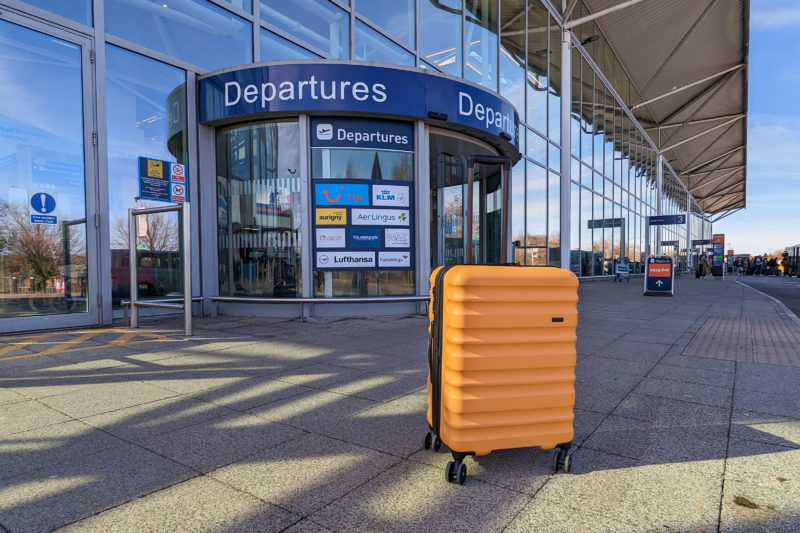 mustard yellow hard shell suitcase with a black handle on concrete floor outside a glass fronted airport building with a blue departures sign above the door