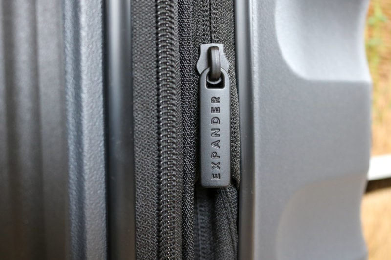 close up of a black zip with the word EXPANDER on it on the side of a dark grey suitcase