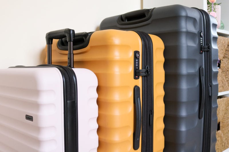 row of three hard shell suitcases in different sizes - the closest is pale pink cabin bag size the middle is mustard yellow medium size and the last is a large dark grey suitcase. all three have black wheels and black trip and handles. they are in a row against a cream wall