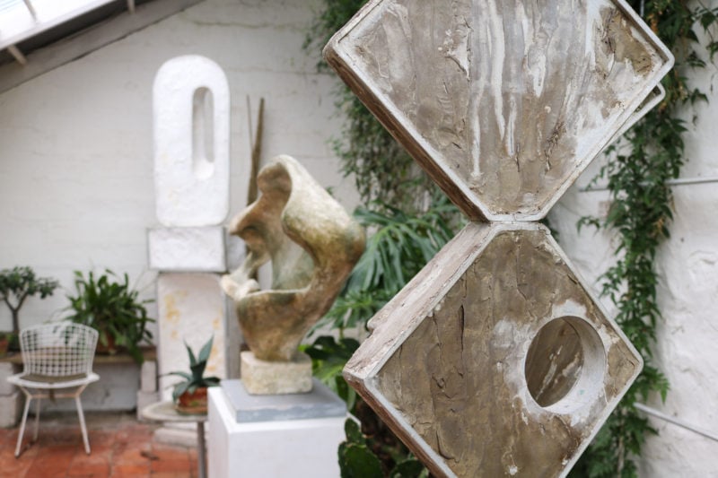 inside the barbara hepworth sculpture museum - interior of a small white conservatory with whitewashed walls and a few sculptures, the closest is made of two large grey diamonds on top of each other, the others are out of focus behind along with some green plants
