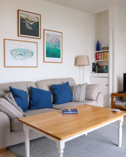 interior of a lounge with white walls and grey carpet with pictures on the walls above a large grey sofa with blue square cushions and a rectangular pine coffee table in front.