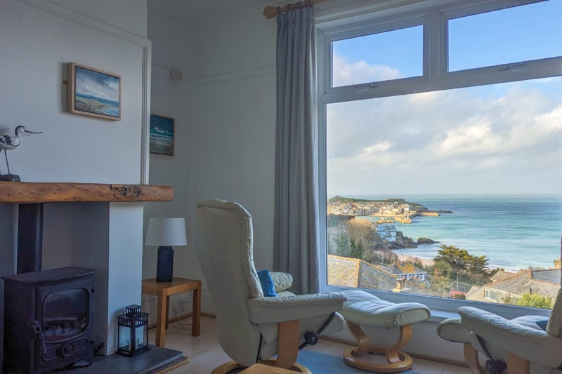 interior of a sitting room with white walls and a small black fireplace with wooden mantlepiece and two cream leather armchairs facing a large window with a view of st ives and the blue sea outside on a very sunny day with blue sky and fluffy white clouds
