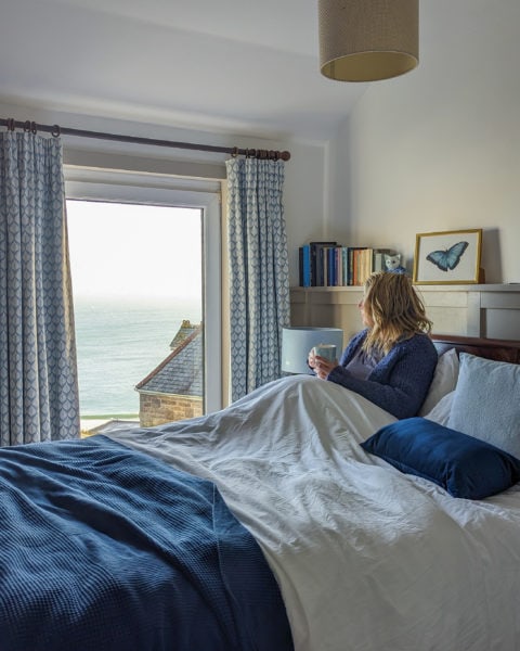 emily wearing a grey top and royal blue woolen cardi holding a coffee cop and sitting in a bed with white sheets and a dark blue throw in a bedroom with white slanted walls and pale blue curtains. the window on the far side of the bed has a view of a grey roof and the blue sea on an overcast day.