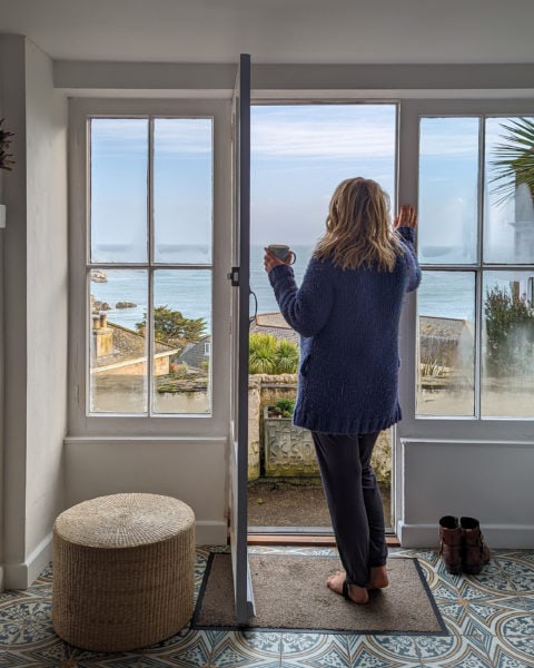 Emily facing away from camera next to a set of white wooden french doors. emily is wearing grey trousers and a long blue woolden cardigan with her long blonde hair loose and is holding a mug of coffee whilst looking out at the view of the sea from bay hill cottage in st ives cornwall england