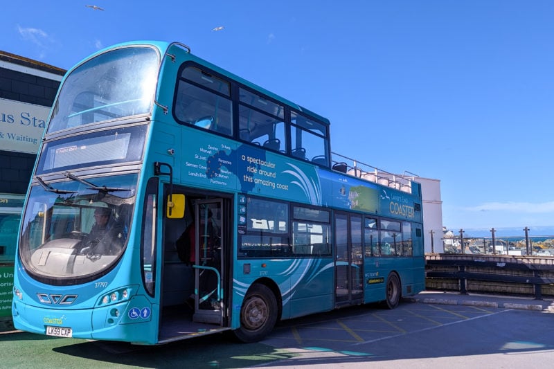 bright blue double decker open top bus with the name Lands End Coaster painted on the side parked in a car park on a very sunny day with clear blue sky above