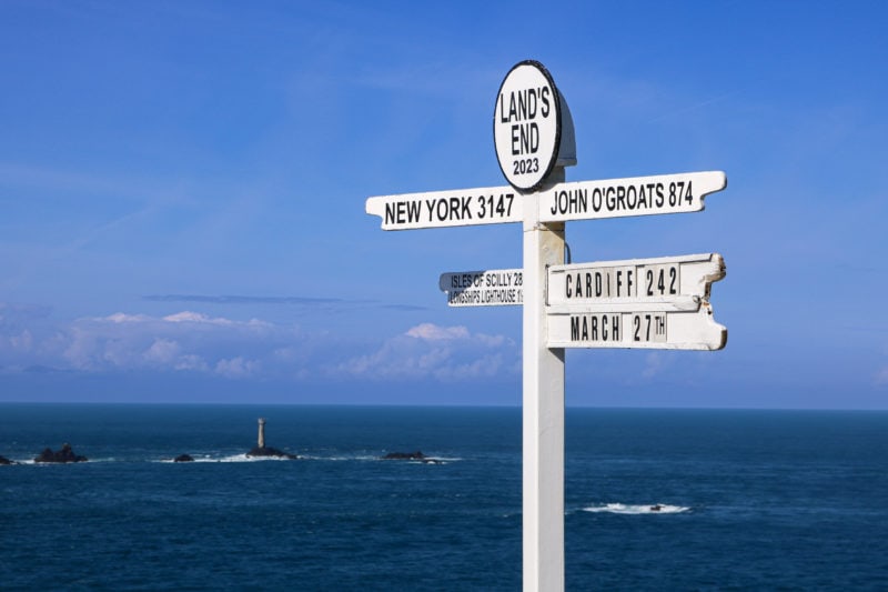 white wooden sign post with Lands End printed in black at the top and finger posts pointing to New York to the left and John O Groats and Cardiff to the right with blue sea and blue sky behind