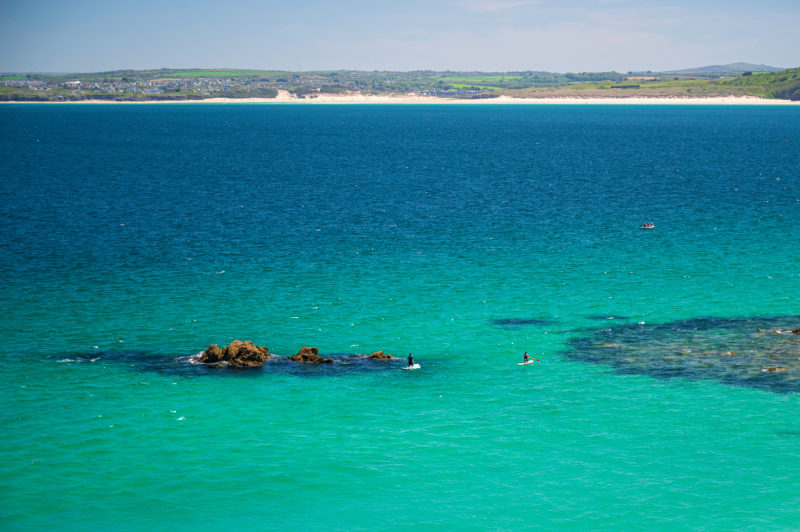 drone shot of very clear turquoise sea water in a large bay with the low grassy cliffs and sandy beach visible in the far distance and two very small paddleboarders next to some brown rocks in the ocean