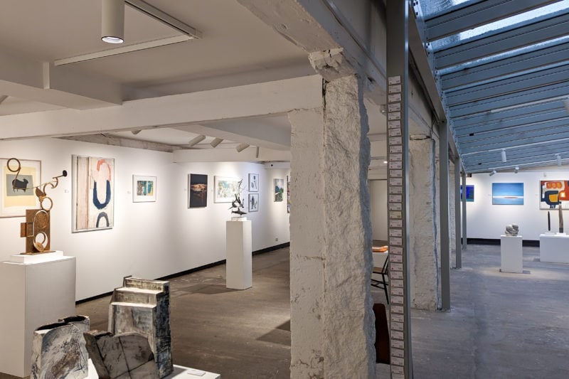 interior of an art gallery with a slanted roof, grey stone floor, white walls, and lots of paintings on the far wall. there are white painted pillars running through the middle of the gallery and a row of slanted skylights in the cieling to the right. 