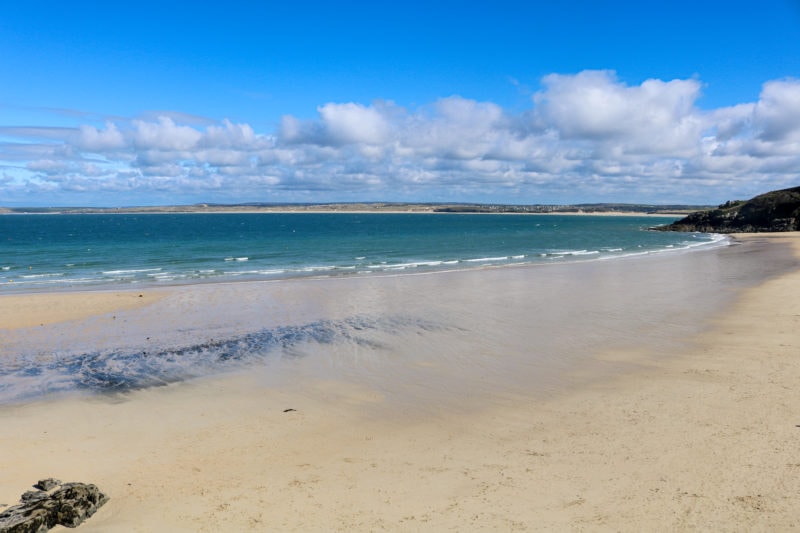 large empty beach with white sand at low tide with the blue sea behind and the grassy far side of the bay visible across the sea in the far distance