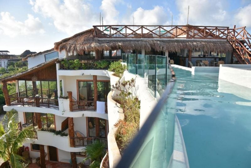 top of a white hotel building with a rooftop pool and a wooden bridge crossing the pool on a sunny day with blue sky and white  clouds 