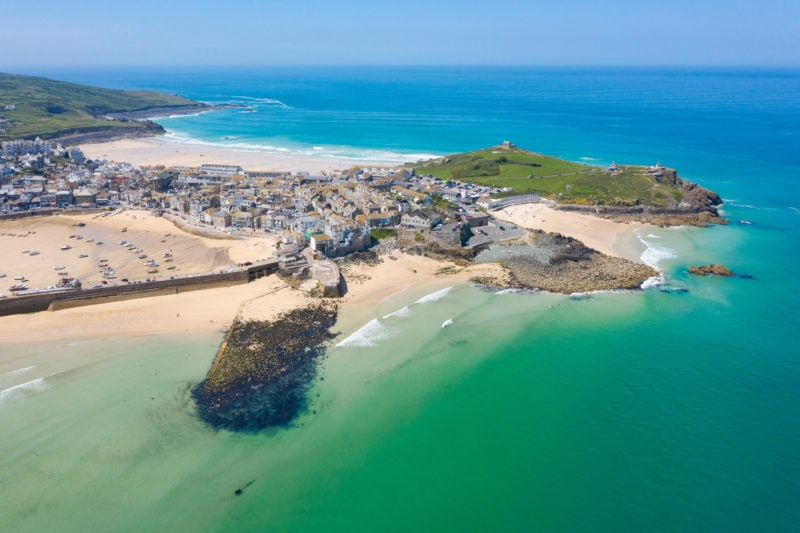 aerial shot of a low headland surrounded nby turquoise sea with golden sandy beaches and a small town with a harbour. What to do in St Ives Cornwall England. 