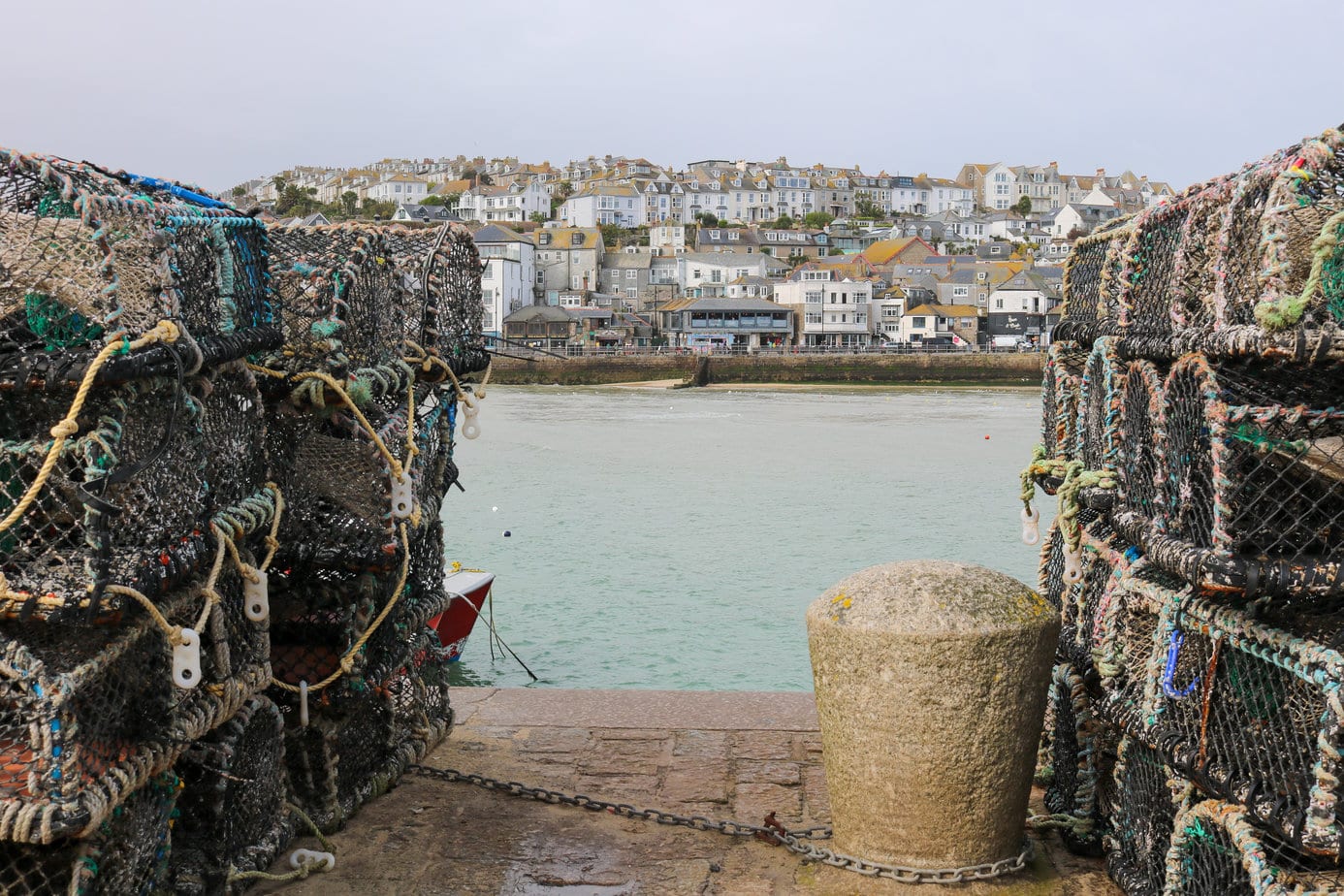 looking between two large stacks of netted fishing cages at a harbour on a grey day with a small town on a hill on the far side - what to do in st ives cornwall england 