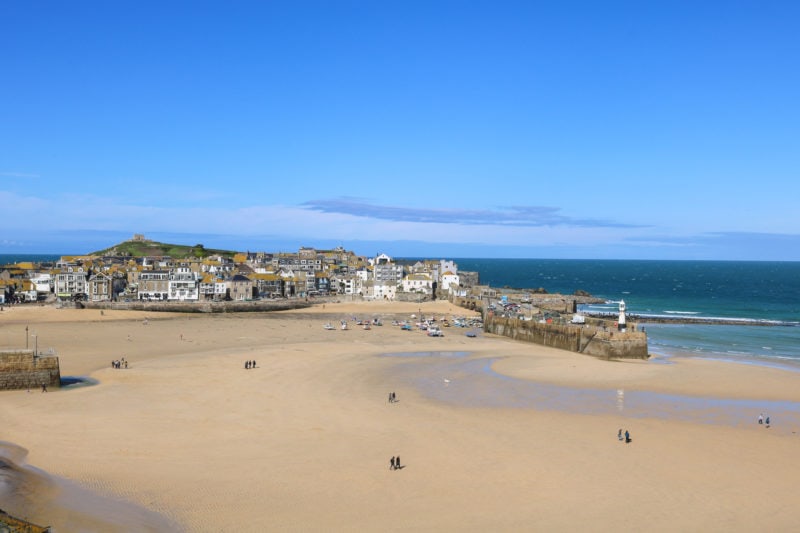 st ives harbour at low tide with the golden sandy floor exposed and a few boats left on the sand. the town and grassy headland are visible behind and an empty blue sky overhead. 