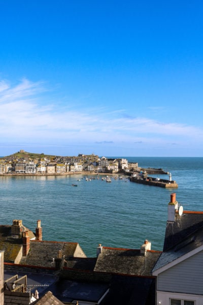 looking past some rooftop chimneys at a large blue harbour protected by a small harbour wall with some small boats inside and the town of st ives and a grassy hill on the other side