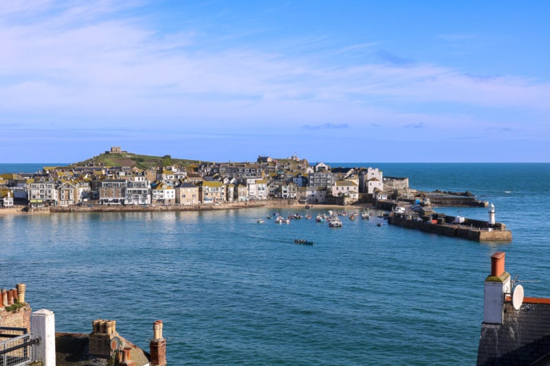 looking past some rooftop chimneys at a large blue harbour protected by a small harbour wall with some small boats inside and the town of st ives and a grassy hill on the other side