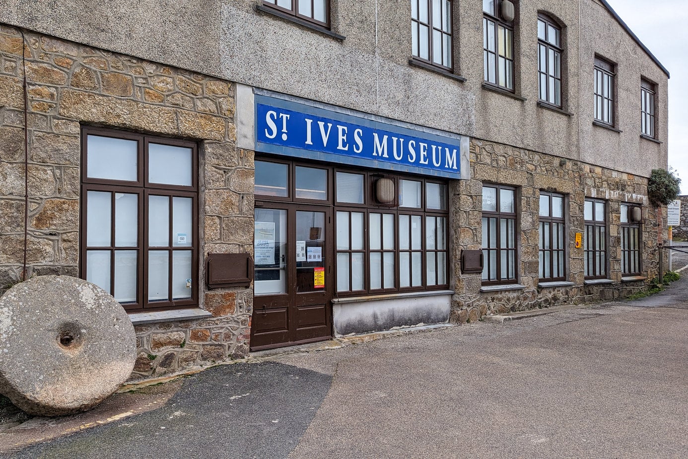 esterior of a large brown and grey stone building with brown frames aroudn the windows and doors and a blue sign that reads St Ives Museum