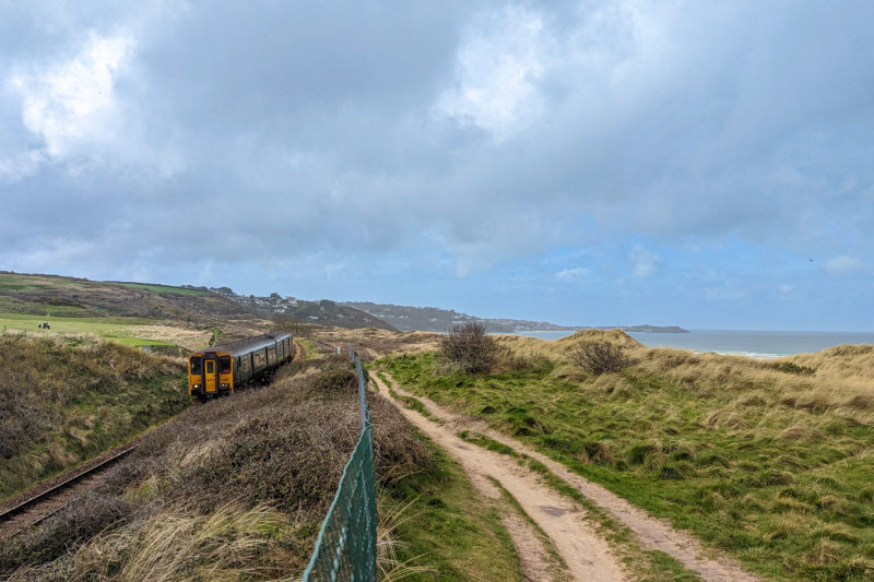 yellow and green train on a railway track to the left of a sandy path running over grassy dunes with a hazy sky overhead 