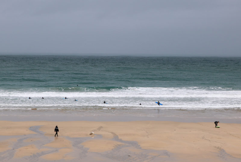 nearly empty sandy beach on agrey day with grey-blue sea behind and some surfers on very small white capped waves in st ives cornwall