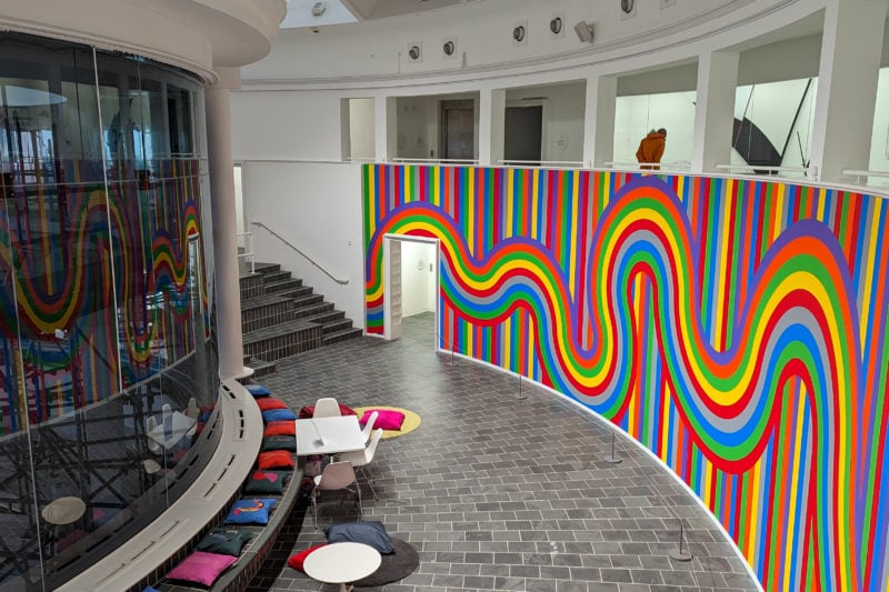 Interior of the Tate art gallery in St Ives with curved wall entirely covered in multicoloured wavy lines and a grey tiled floor with some circular white tables on it next to a large curved window