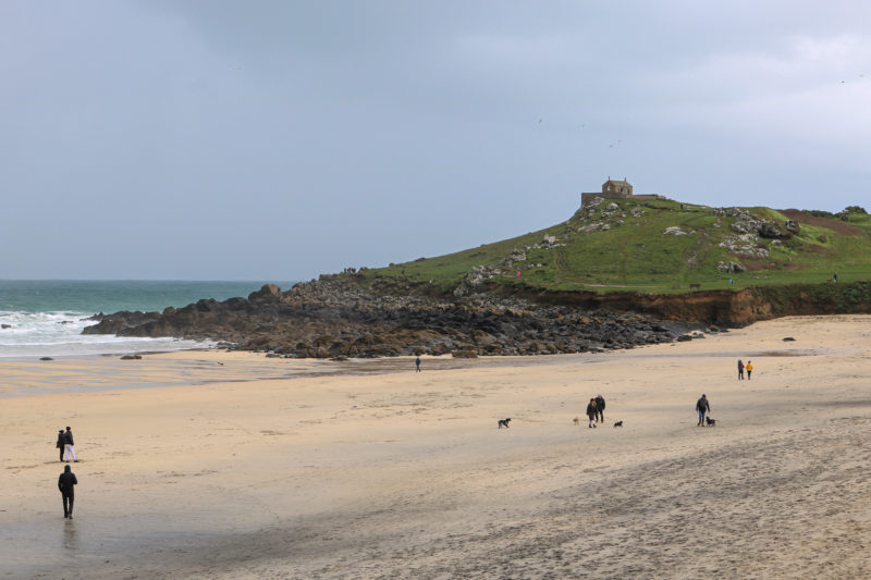 sandy beach on a cold grey day with a few figures walking along the sand and a large grassy hill surrounded by rocks overlooking the sand. there is a small stone chapel on top of the headland. what to do in st ives cornwall england