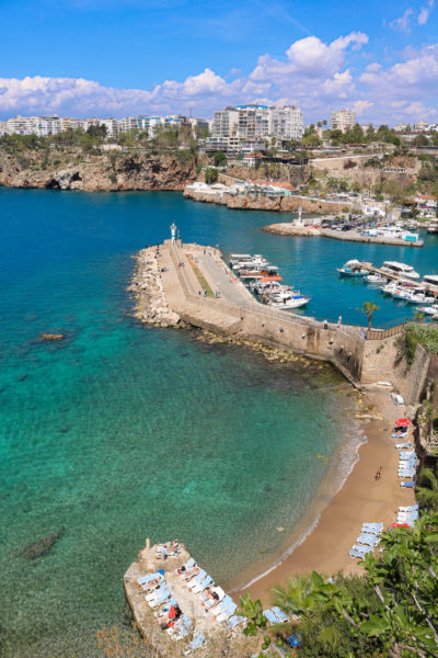 looking down from a cliff at a small sandy bay with turquoise sea and bathers on sunbeds on a small stone pier with a longer stone pier on the other side of the bay and a small harbour with fishing boarts behind that and the town of antalya in the distance on a very sunny day with blue sky - Jet2 Treasures of Turkey review