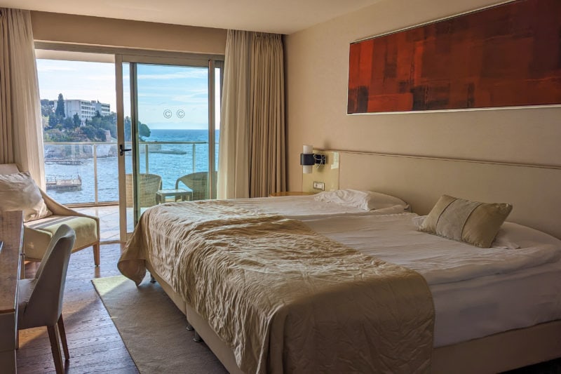 interior of a hotel room with cream walls and two large double beds with white bedding beneath a long rectangular painting of dark red shapes. there are glass sliding doors at the end of the room with a balcony outside and a view of the blue sea and a rocky coastline visible through the window. hotel room at Charisma Deluxe Hotel in Kusadasi Turkey.