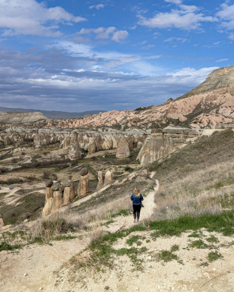 emily wearing black trousers and a dark blue denim jacket walking down a sandy path away from the camera with a large dry valley beyond filled with tall conical rock stacks. The floor and rocks in the valley are yellow sandy coloured stone with small patches of grass growing here and there and blue cloudy sky overhead. 