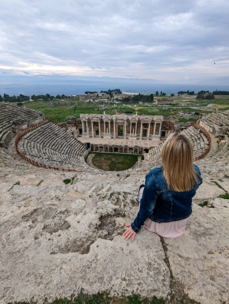 Emily wearing a dark blue denim jacket with her long blonde hair loose facing away from the camera sitting at the top of stone steps which are layed out in semi circular rows in Hieropolis Ancient Theatre in Turkey on an overcast day with light grey cloudy sky above. 