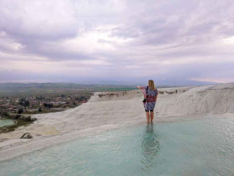 emily wearing an orange and white patterned shawl and standing on the edge of a light blue pool of water framed with white rocky edging formed by mineral deposits with a white cliff sloping away behind her and a green landscape in the distance below on an overcast evening with dark grey clouds above