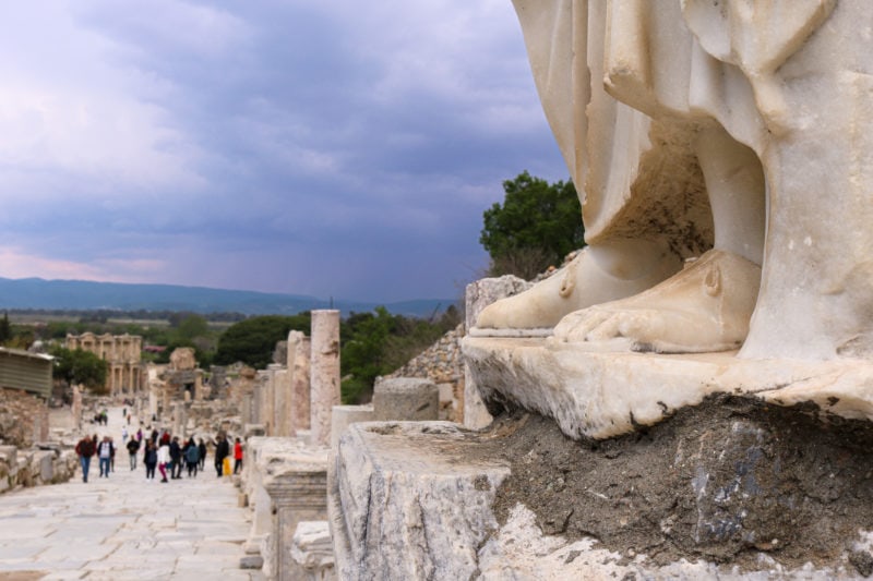 close up of the feet of a white marble statue - bare human feet on top of a grey pillar with a white stone road and distance ruins of a city out of focus in the background and a grey sky overhead