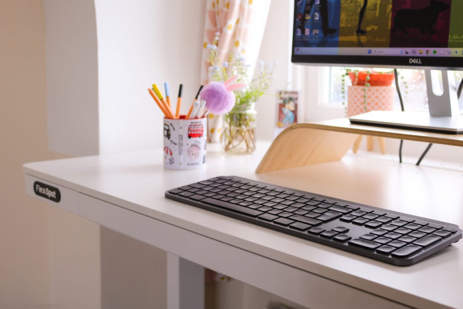 corner of a white wooden desk top with a black keyboard, a mug of pens and the lower half of a monitor visible. FlexiSpot Comhar EW8 review.