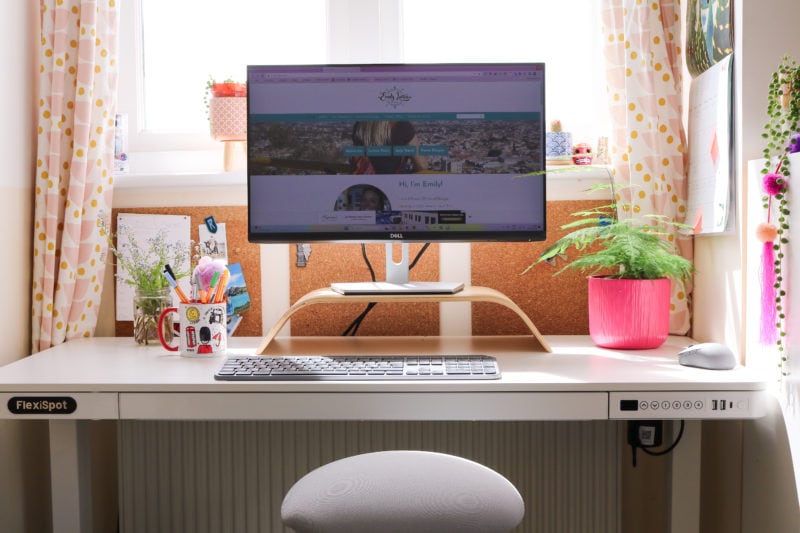 white desk in an alcove under a window with a computer monitor on it on top of a wooden stand. there is also a pink plant pot with a fern, a glass jar with forget me not flowers in it, and a mug full of pens on the desk. there is the top of a grey stool with a cushioned seat visible in front of the desk.
