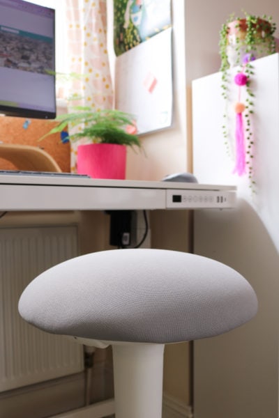 close up of the top of a stool with a padded grey cushioned seat and cream coloured stand in front of a white desk and bookshelf which are out of focus.