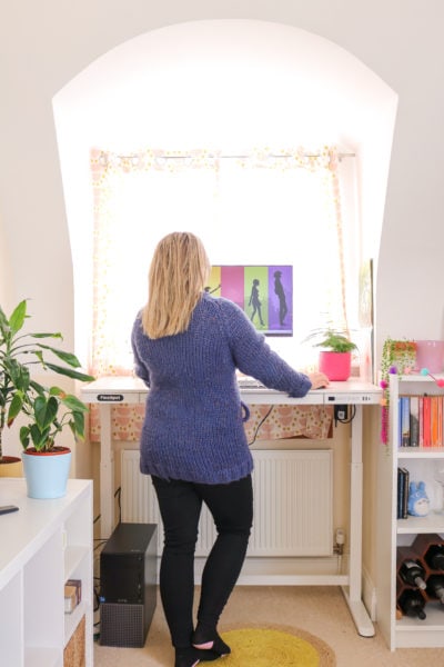 emily wearing black jeans and a long blue woolen cardigan with her long blonde hair loose standing in front of a white desk with a monitor on it facing away from the camera. there is a window behind the desk.