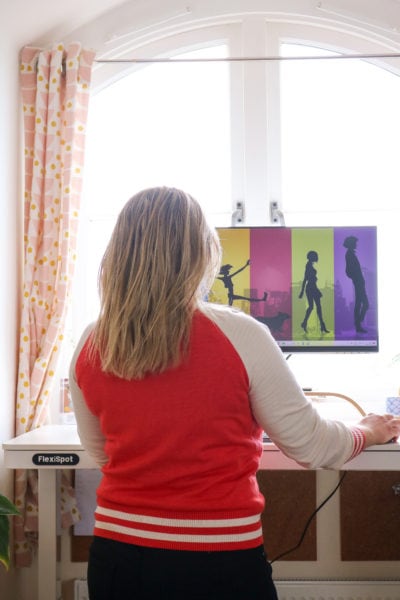 emily wearing black jeans and a red and white sweater with her long blonde hair loose standing in front of a white desk with a monitor on it facing away from the camera. there is a window behind the desk with pale pink curtains. flexispot comhar ew8 standing desk review. 