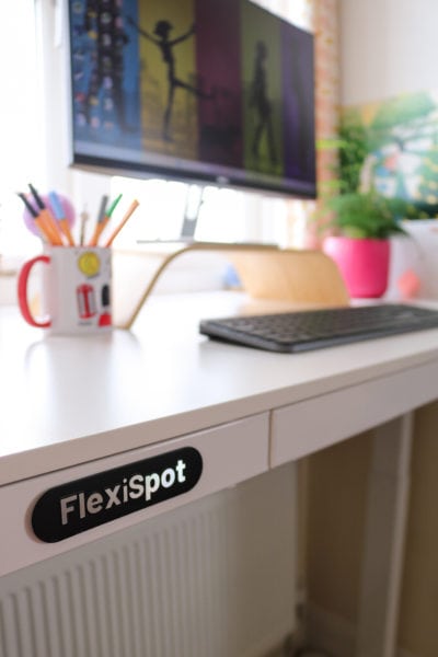 close up of the edge of a white wooden desk top with the word Flexispot in silver on a black oval on the side of the desk. there is a pc monitor on the desk with a black keyboard in front and a mug full of pens, and also a pink plant pot with a fern out of focus on the far side of the desk.