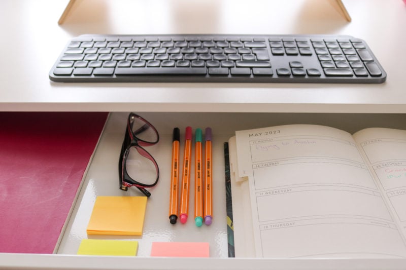 close up of a white wooden desk top with a black keyboard on it, and a very slim desk drawer open in the front. Inside the drawer is a pink notebook, a pair of pink framed glasses, some pink and yellow post it notes, four orange pens, and an open diary.